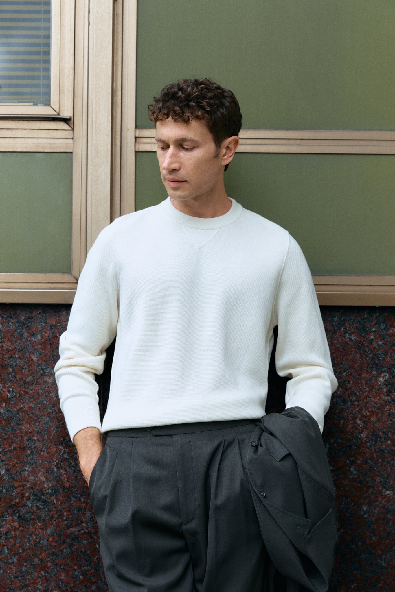 Man wearing a white wool sweater and dark pants against a green wall.