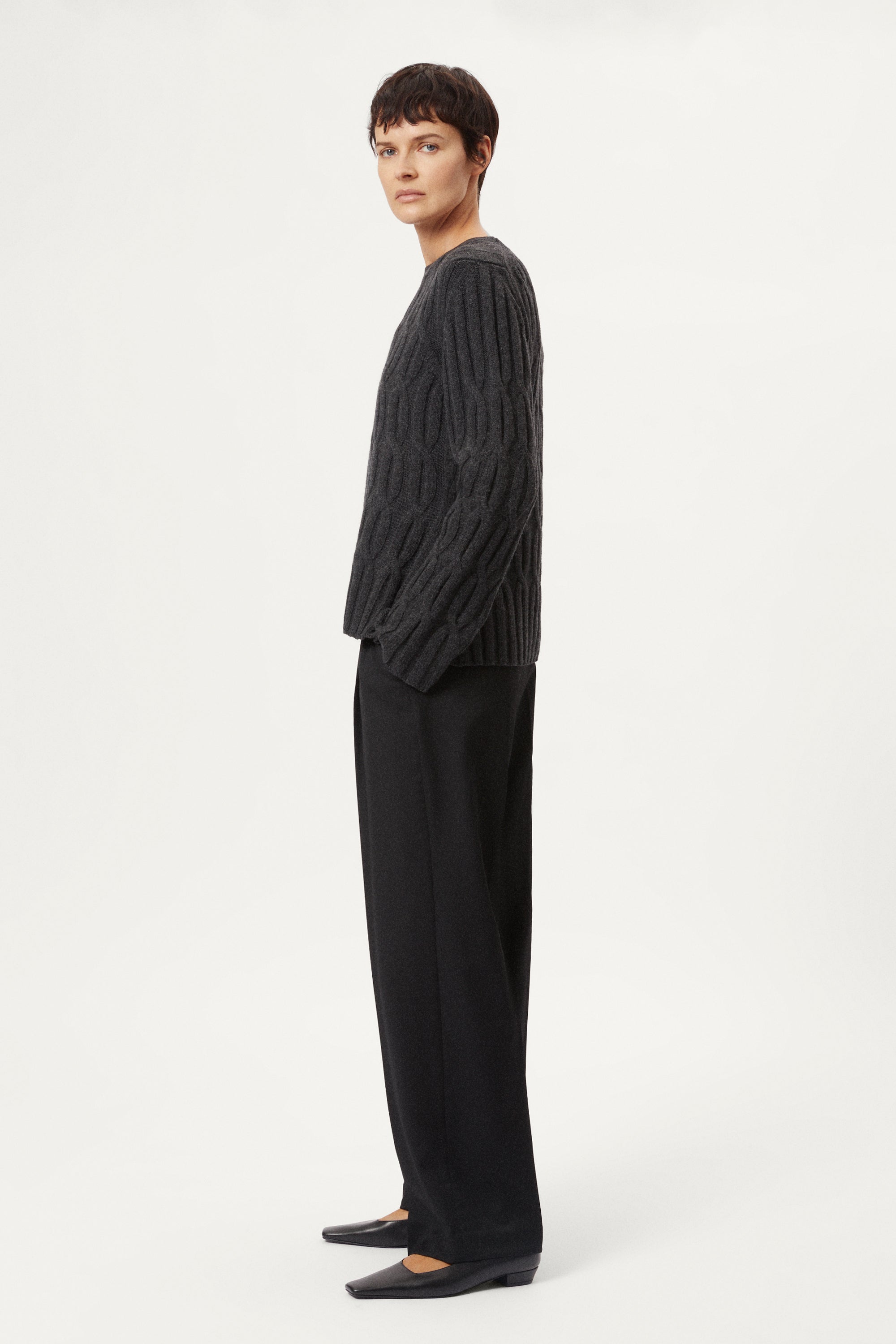 Woman wearing a woolen dark gray cable knit sweater and grey pants on a white background