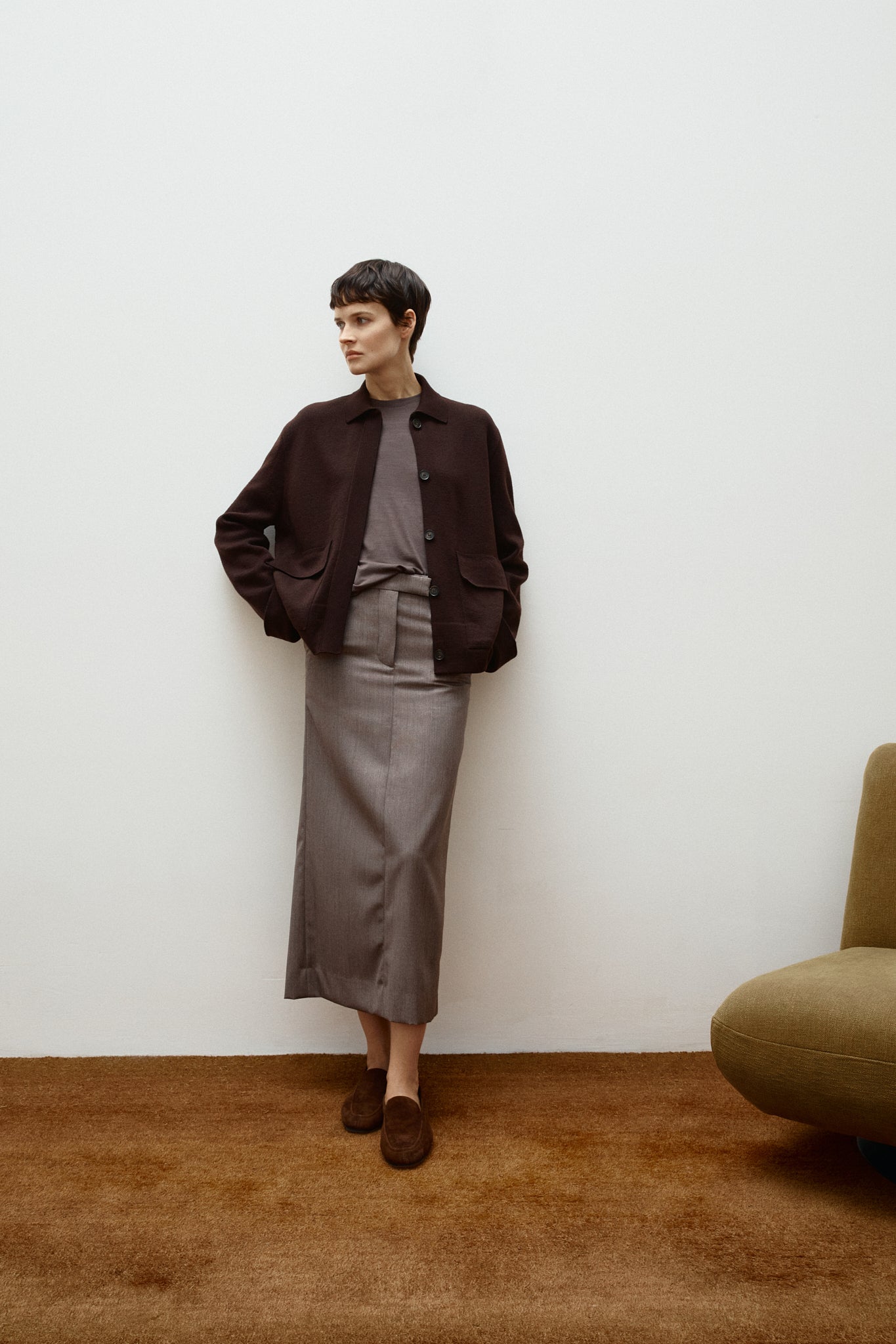 Person wearing a brown wool  jacket and gray skirt standing against a white wall with a brown carpet.