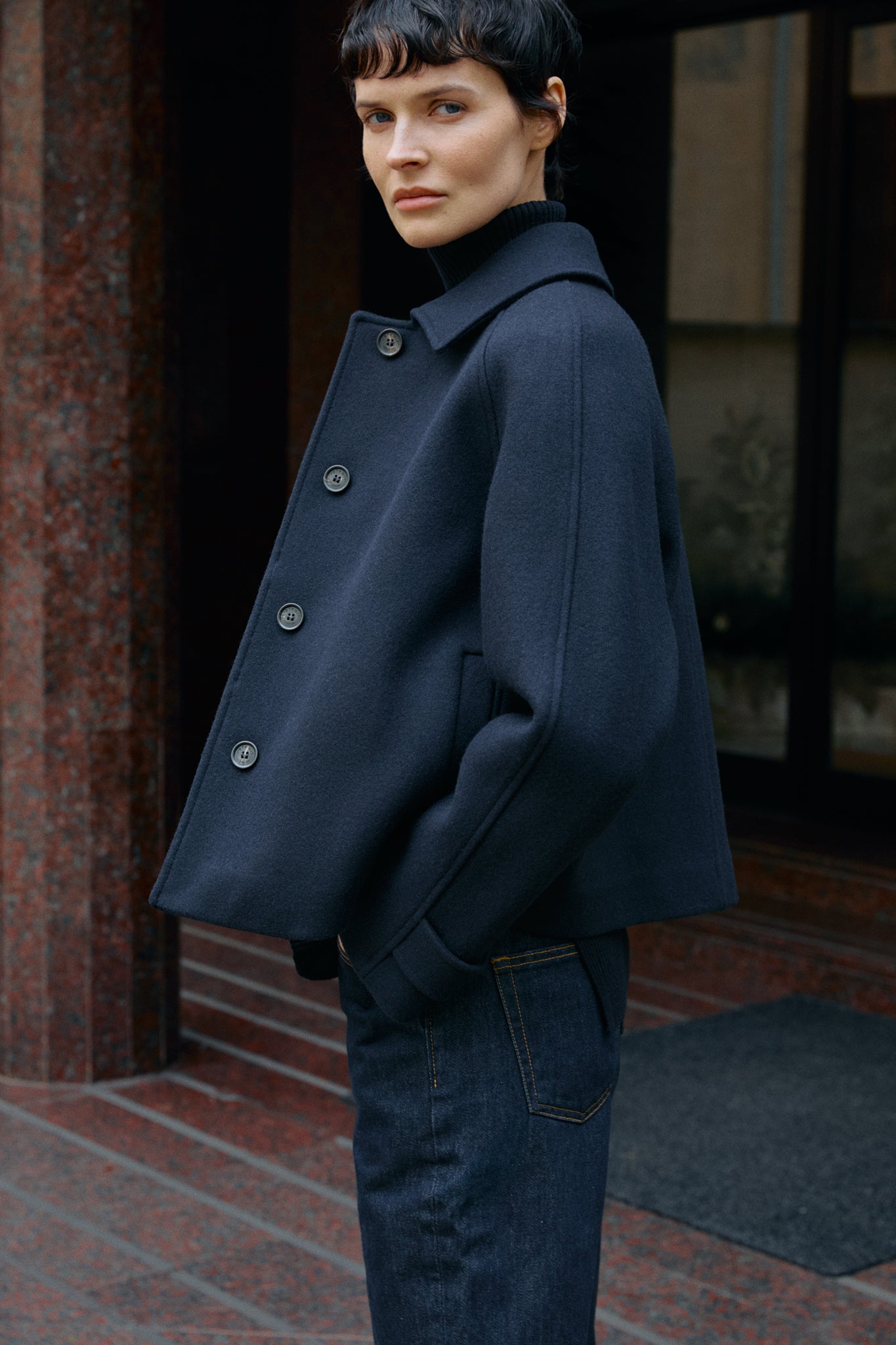 woman wearing a dark blue short coat standing against a building entrance.