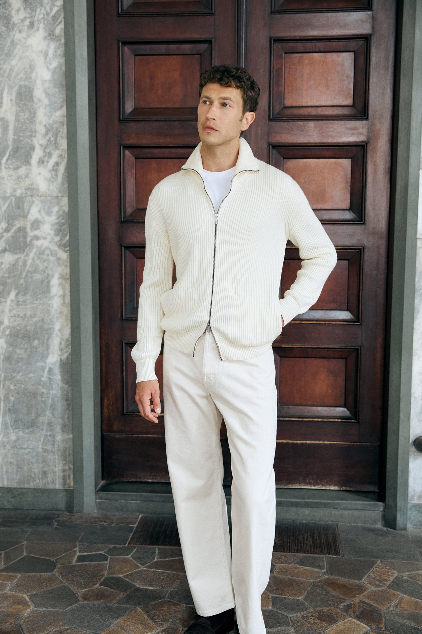 Man wearing a white wool outfit standing in front of a wooden door.