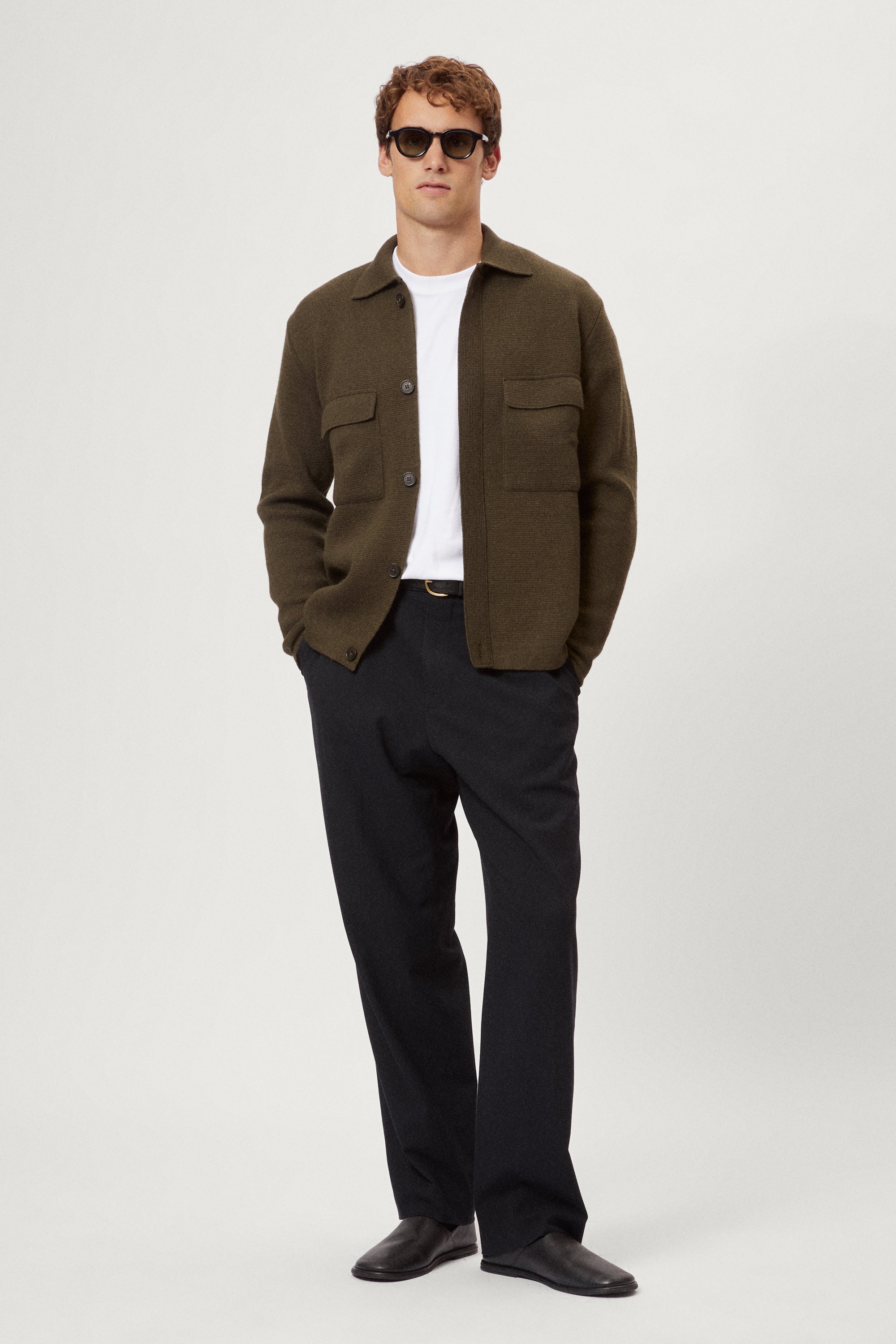 Man wearing a merino green jacket, white t-shirt, black pants, and sunglasses on a white background