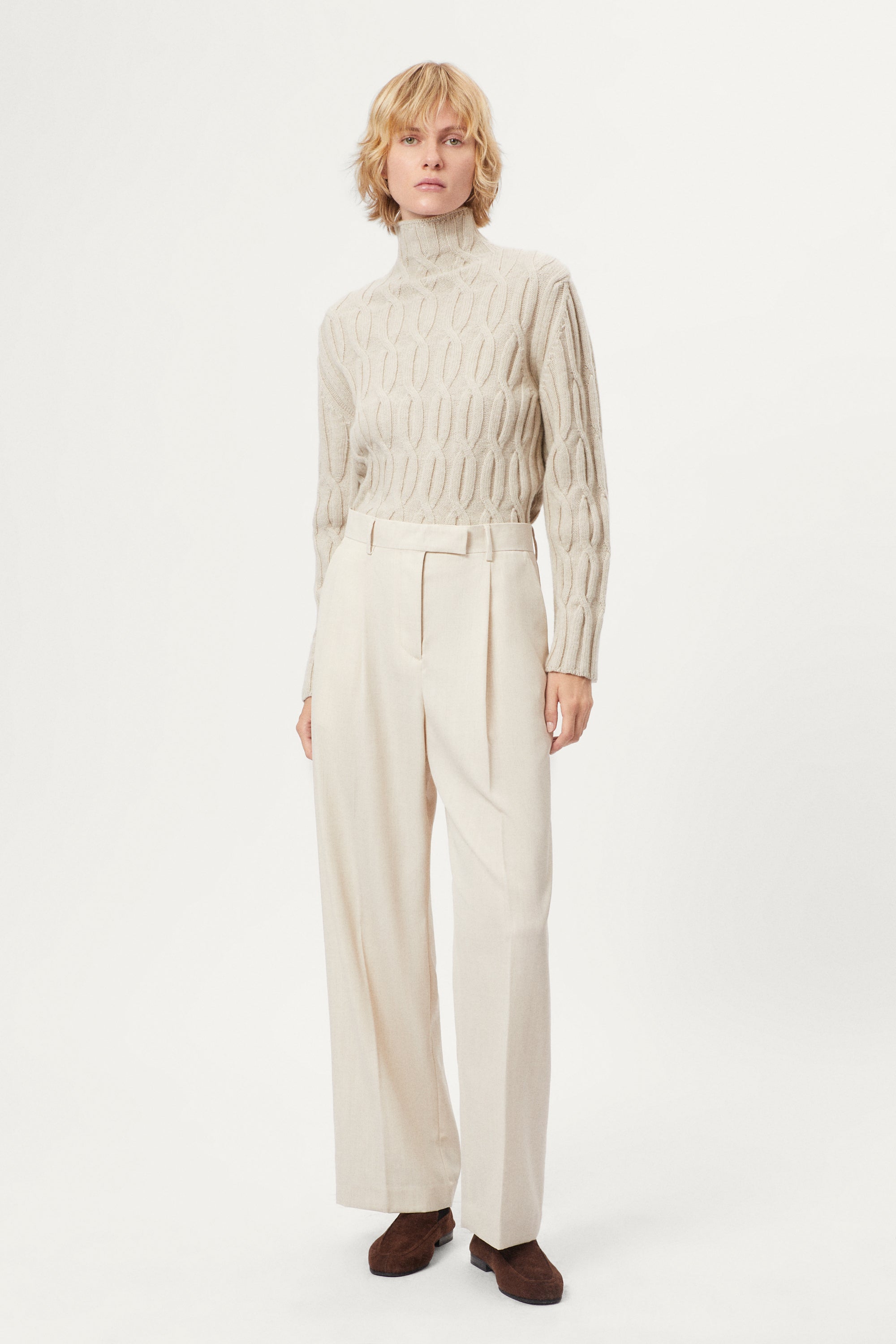 woman wearing a woolen  beige cable knit turtleneck sweater on a white background