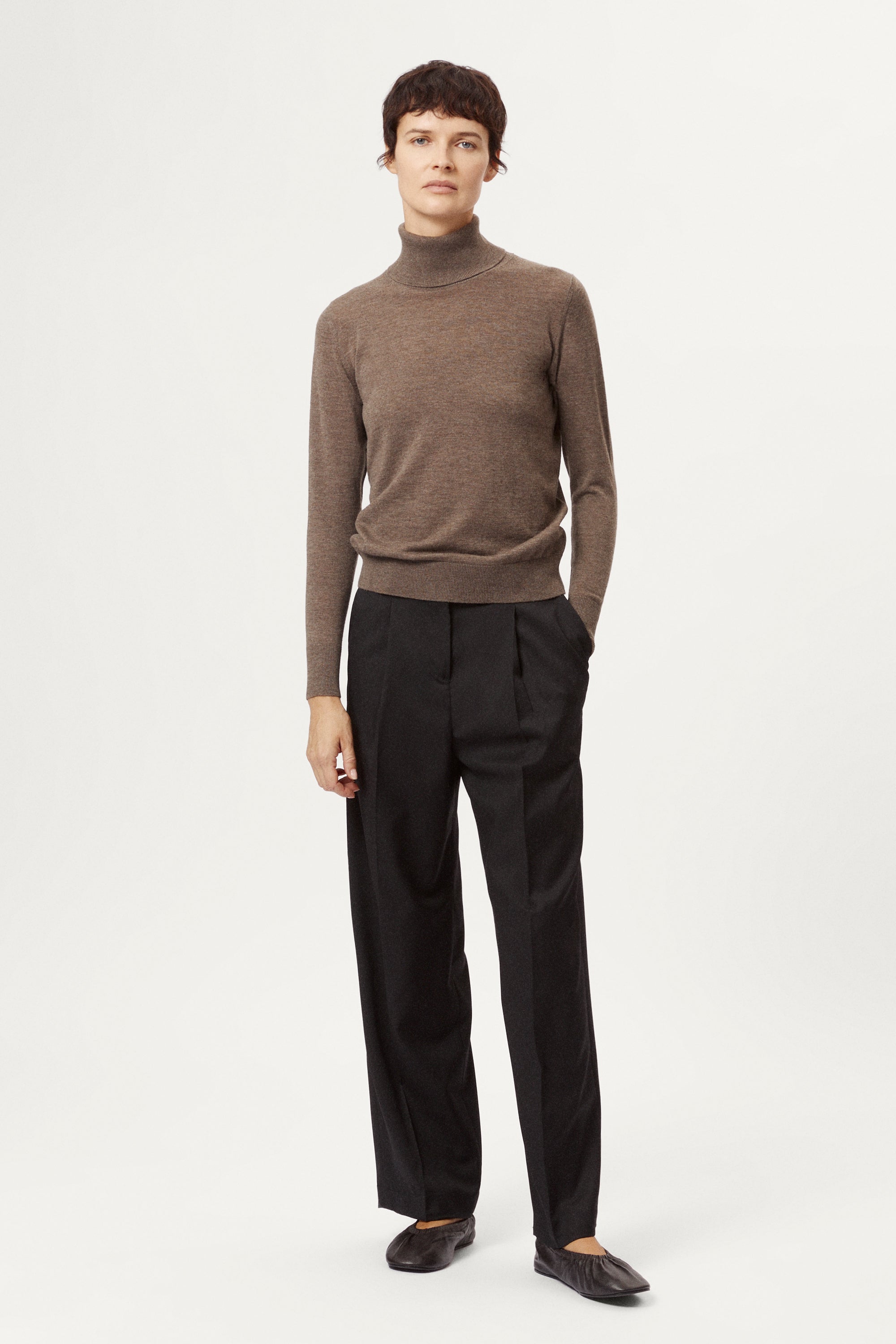 Person wearing a brown turtleneck sweater and black pants on a white background